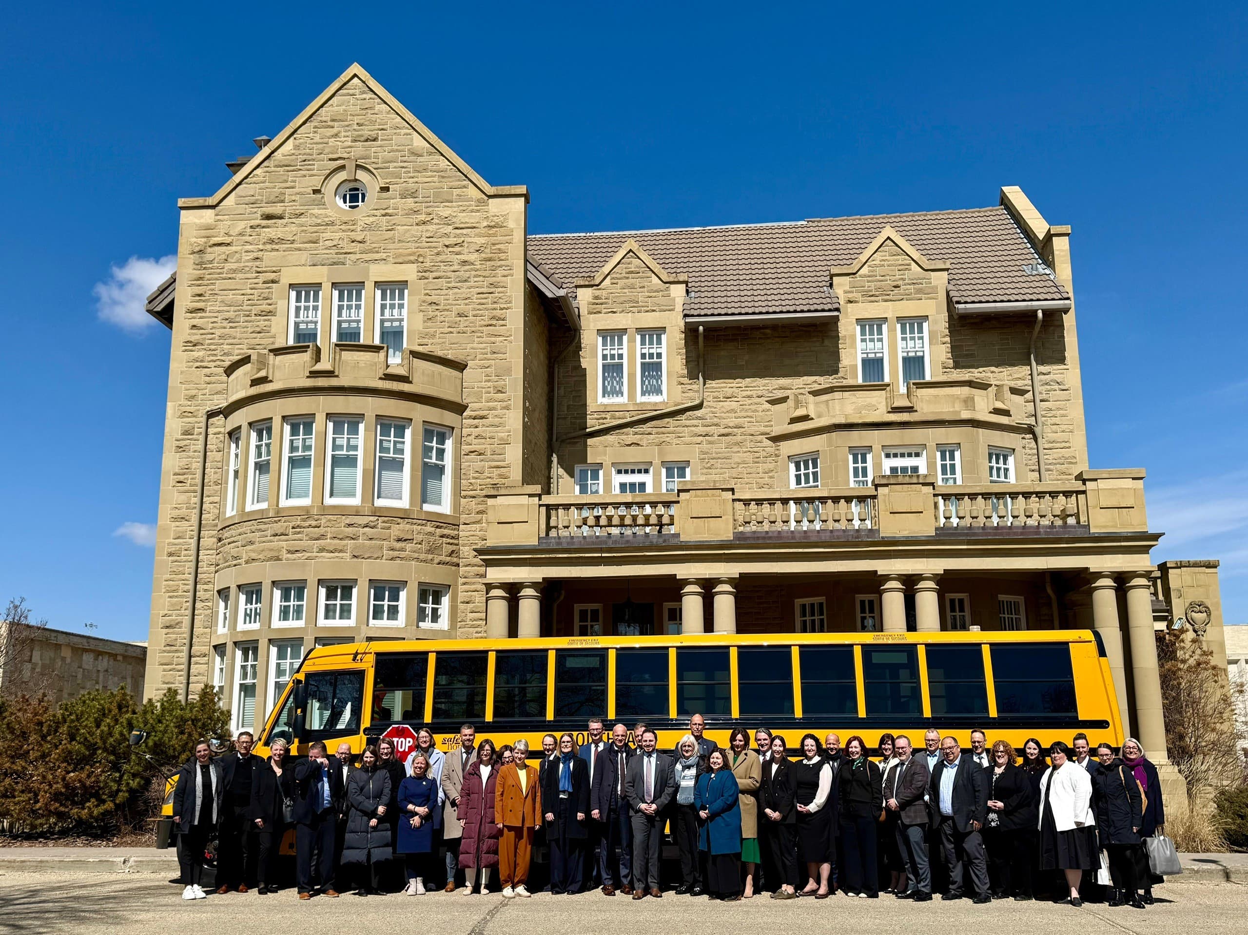 Eine deutsche Delegation steht vor dem Bildungsministerium der kanadischen Provinz Alberta. Die Gruppe posiert vor einem historischen sandsteinfarbenen Gebäude; dahinter ist ein gelber Schulbus zu sehen. Der Himmel ist klar und blau.