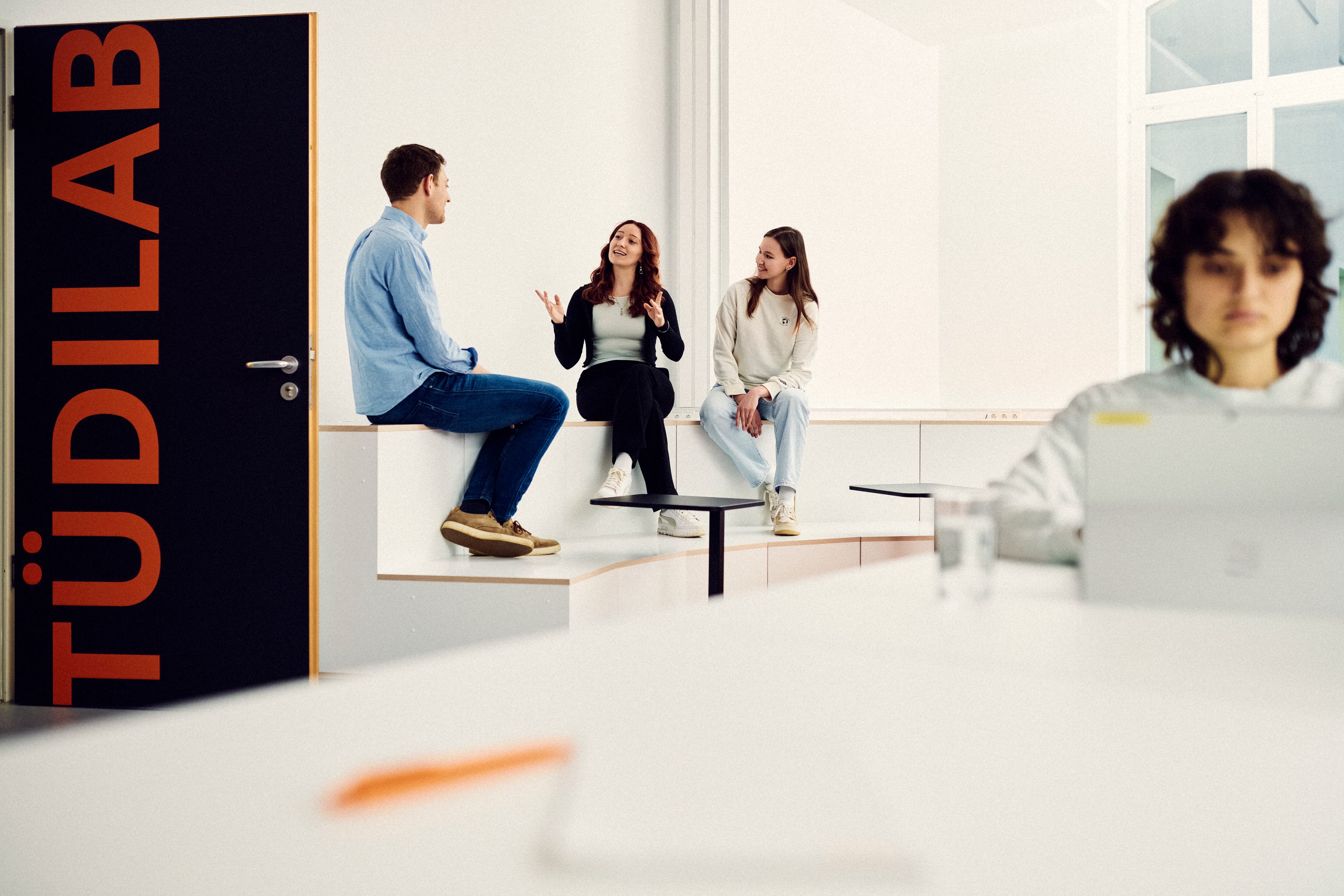 One person working on a laptop in the TüDiLab while three people are talking in the background